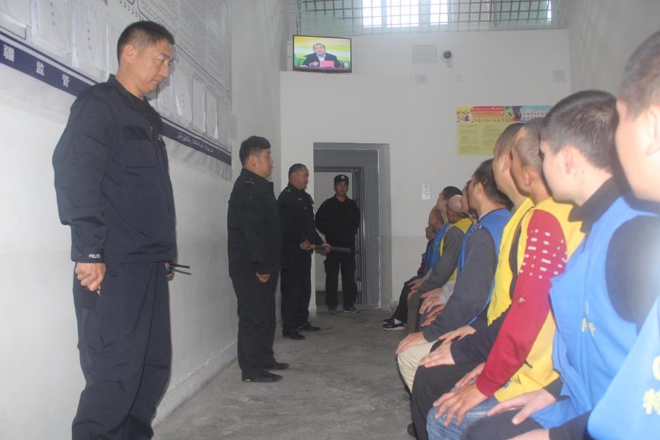 Photo of detainees watching TV while guards stand over them