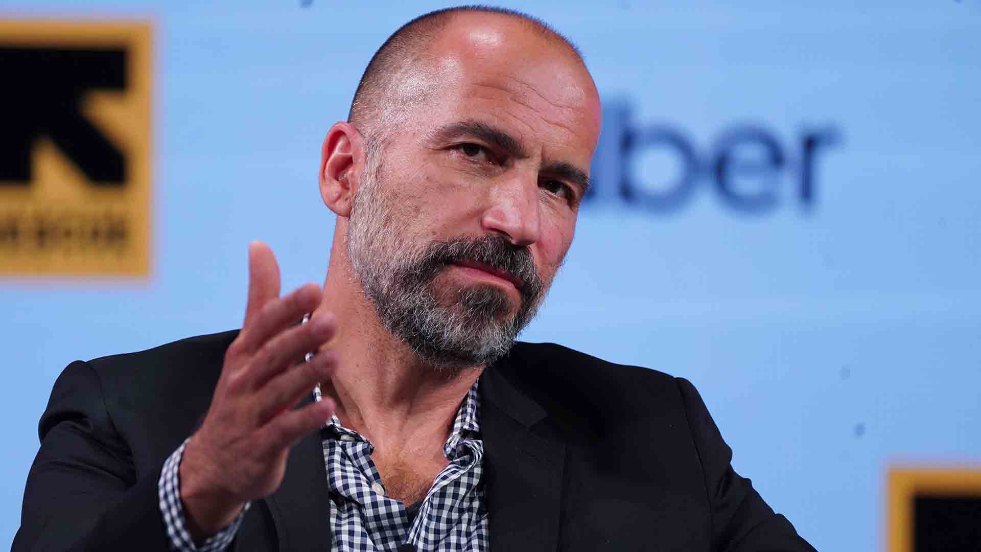 A man with a beard and shaved head raises his hand while speaking in front of a sign that says Uber