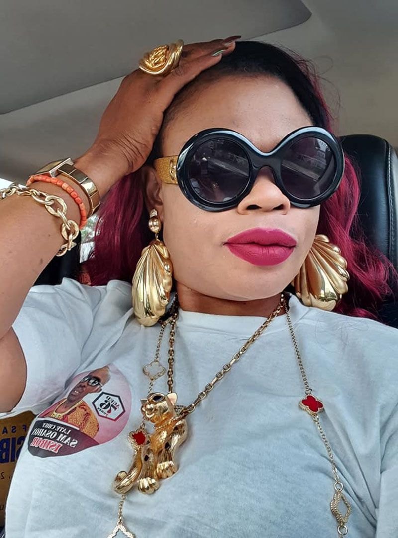 A selfie of a woman with red lipsit, oversized black sunglasses and gold jewelry sitting in a car.