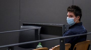 A man in a blue shirt with spiky hair wears a surgical mask while sitting in a courtroom