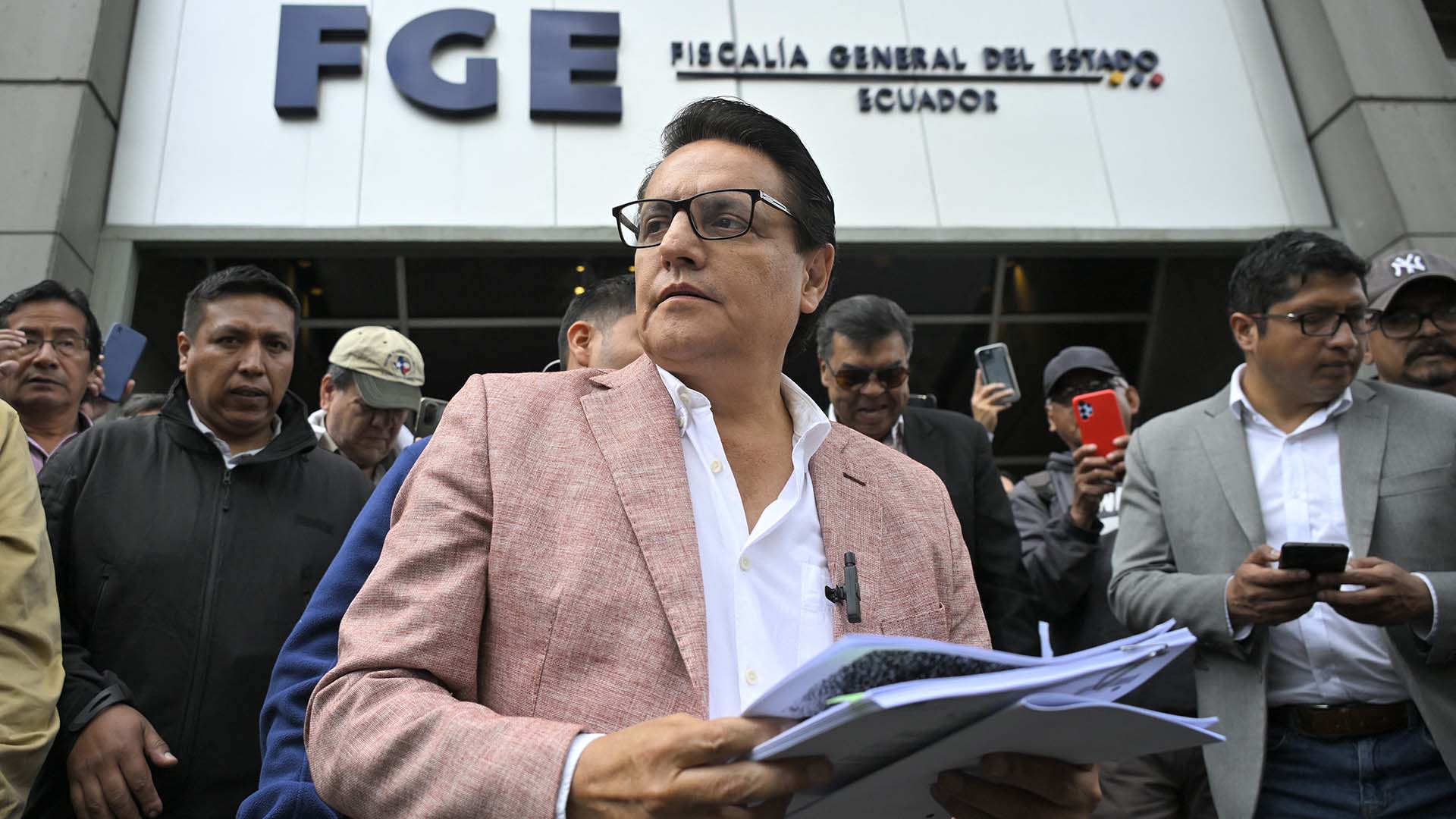 A man in glasses and a pink jacket holding a stack of papers at a press conference.