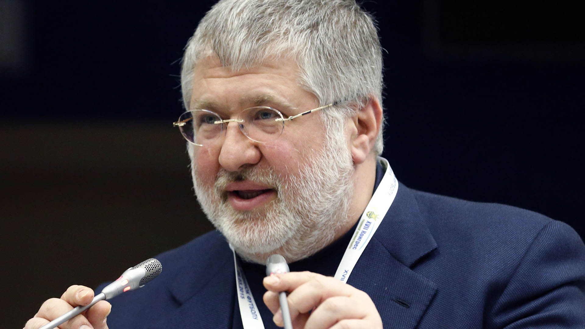 A man with white hair glasses and a beard talks into two small microphones