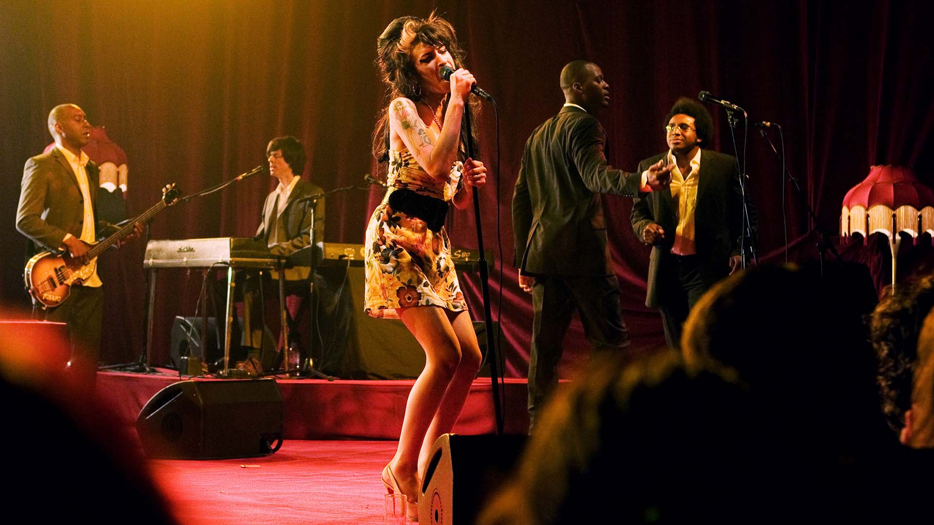 Amy Winehouse singing into a microphone, surrounded by musicians on a stage