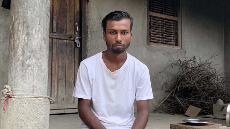 A Nepali man in a white t-shirt sits on the steps of his house, looking in to the camera