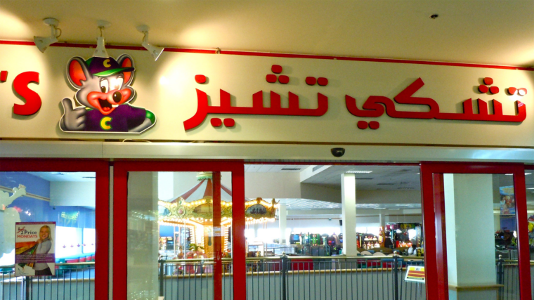 The storefront of a Chuck E. Cheese labelled in Arabic lettering. Through the window, arcade games and a merry-go-round are visible.