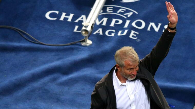 Roman Abramovich holding one hand high to wave, against a blue UEFA Champions League backdrop.