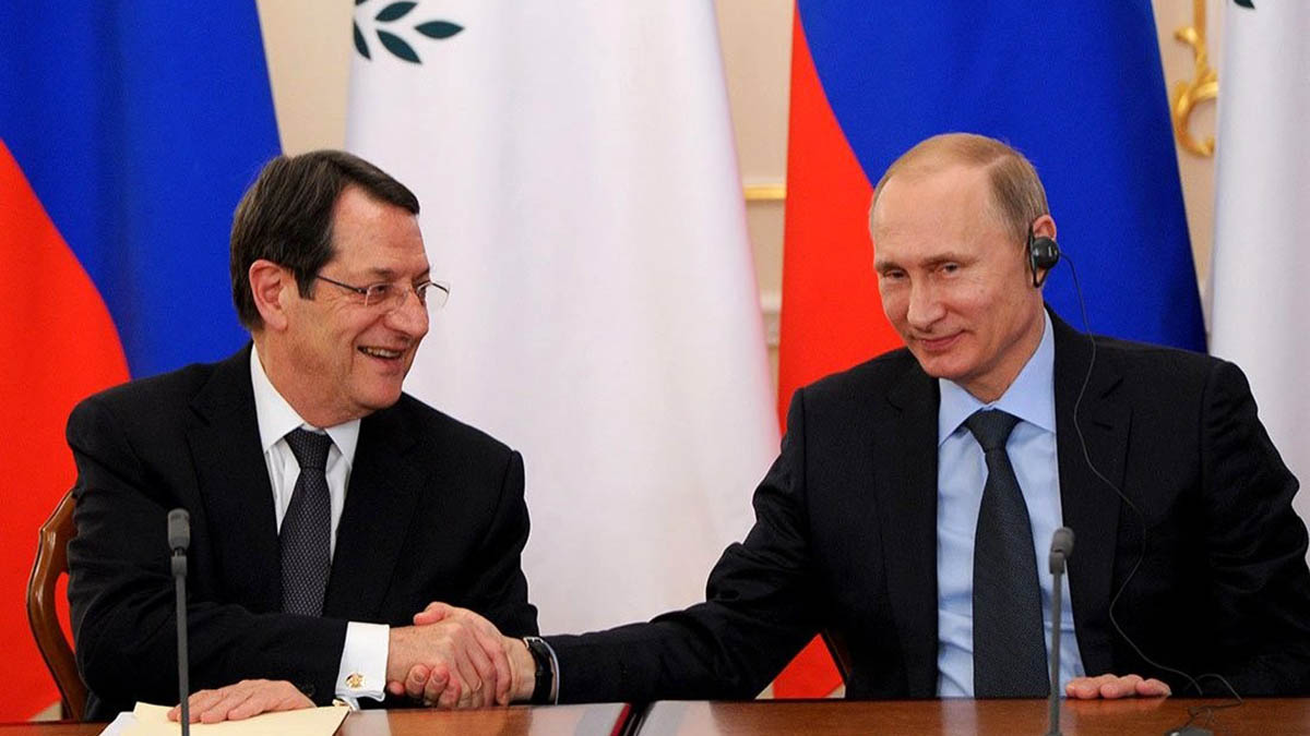 Two men shaking hands while seated at a desk in front of Russian and Cypriot flags.