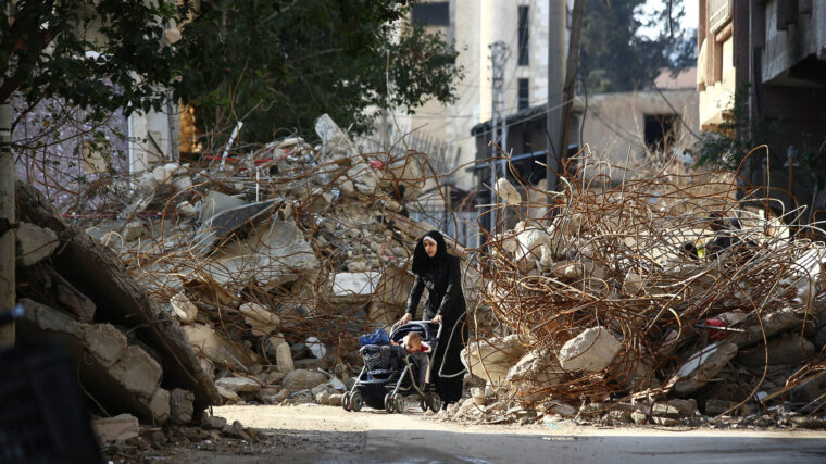 A woman walks pushing a baby in a stroller between the rubble of destroyed buildings.