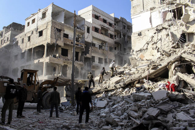 Several people climb upon a hill of white debris surrounded by dilapidated buildings. A construction vehicle sits at the bottom. 