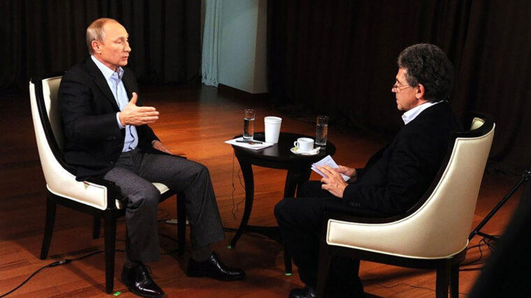 Two men sit opposite each other for a TV interview in white, curved chairs