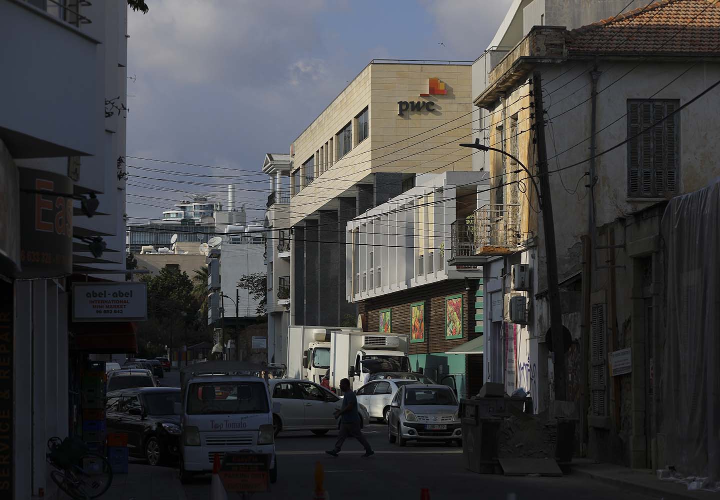 Street scene with cars, a man walking across a road, and a taller building topped with a PwC sign in the background.