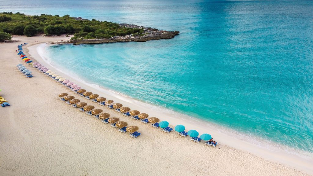 Aerial photo of deck chairs and umbrellas lined up along a sandy beach next to azure waters.