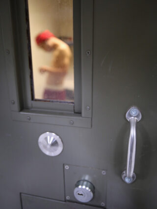 A man in a red hat through the small window of an isolation cell in immigration detention
