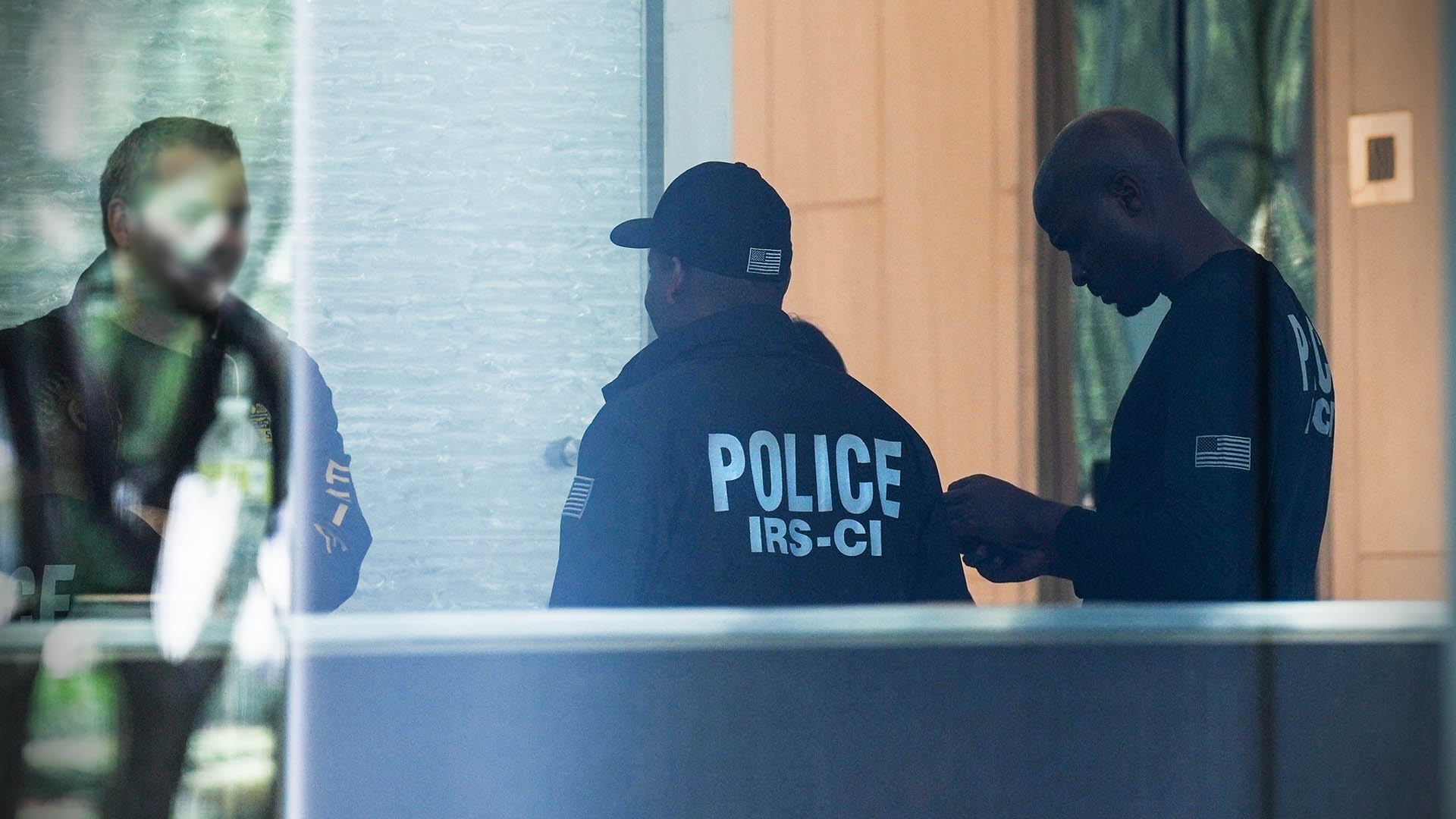 Three men in the lobby of a building, one wearing a jacket that reads POLICE IRS-CI on the back.