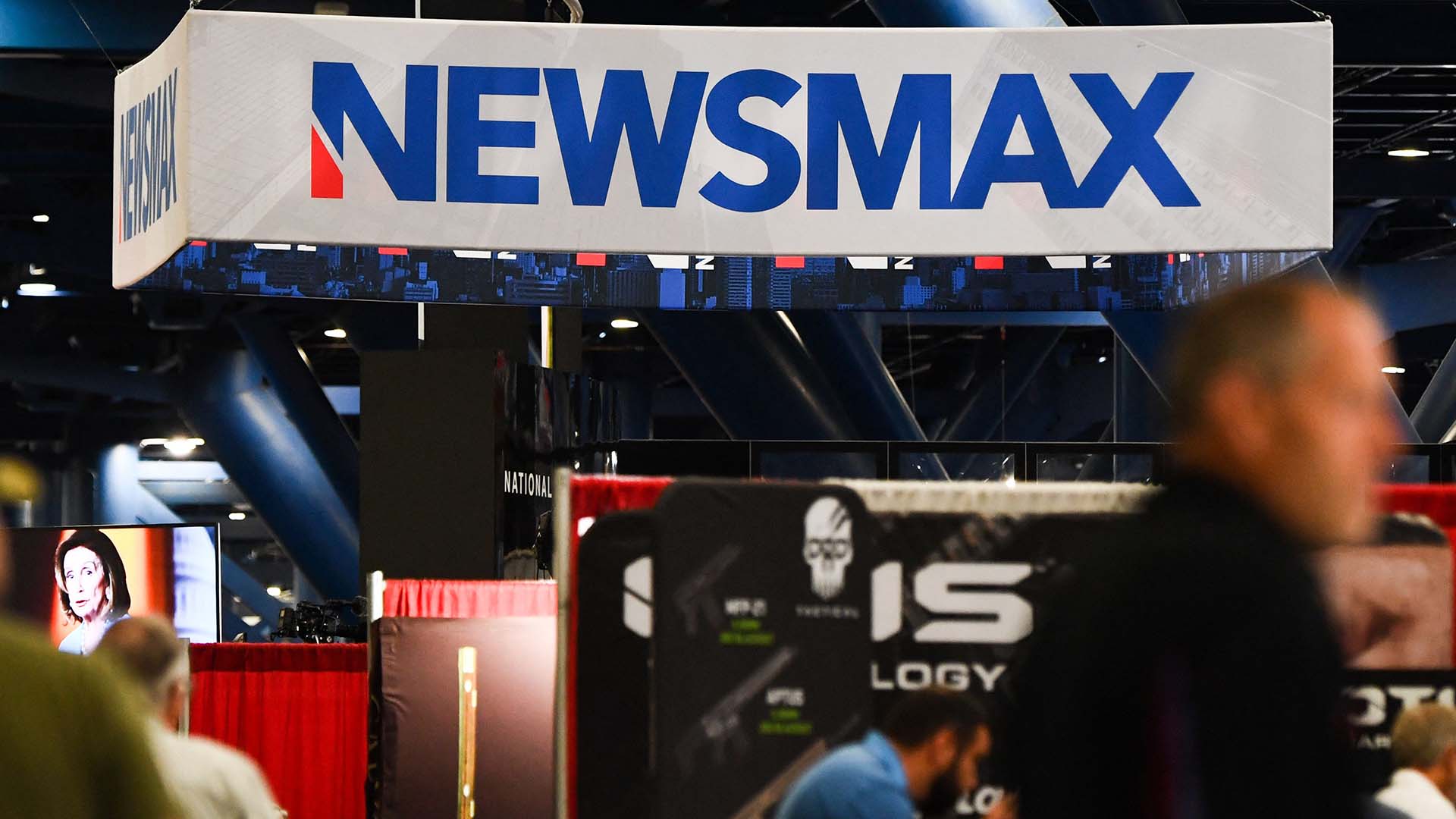 Large sign reading Newsmax above a stall inside a conference hall, with people walking past.