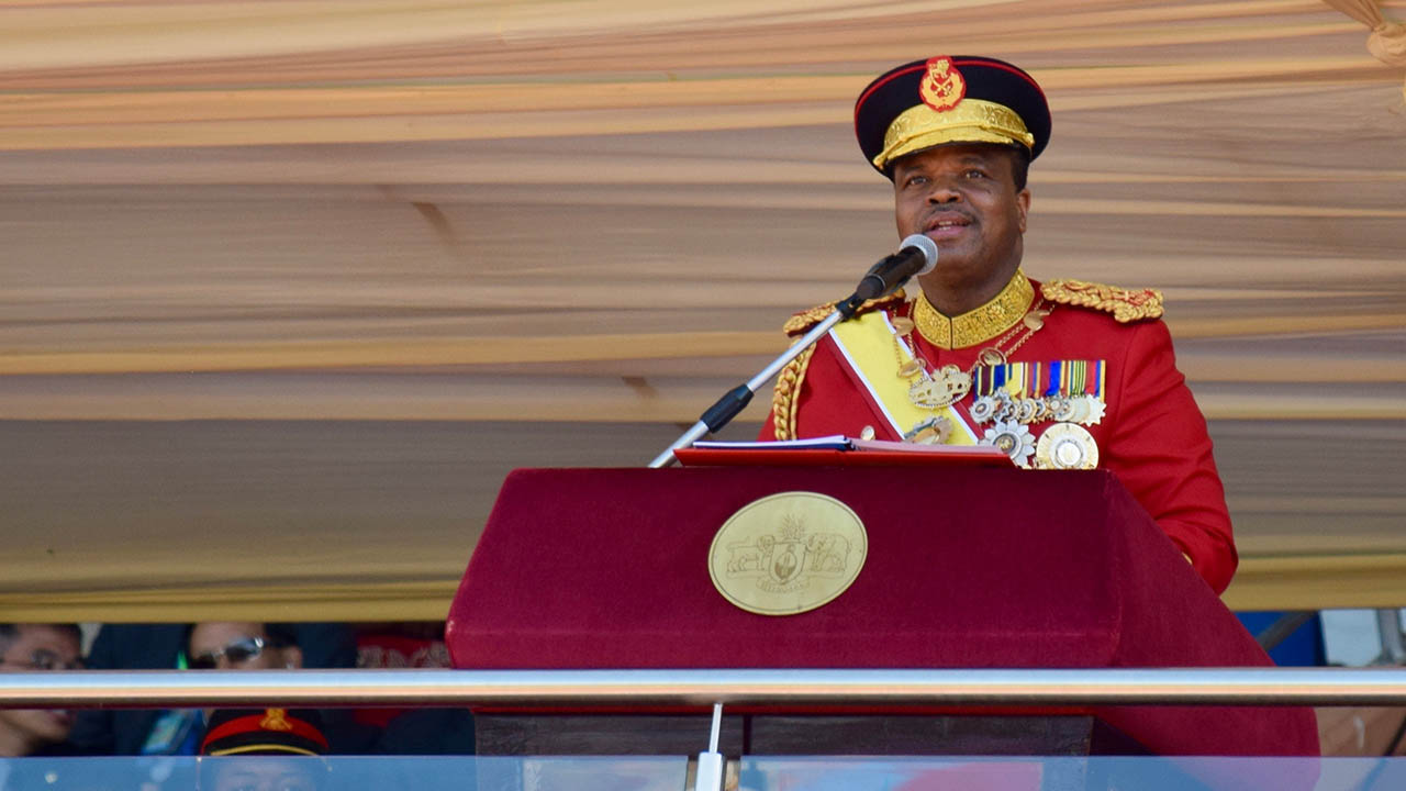 King Mswati III in red military uniform with medals, speaking behind a podium.