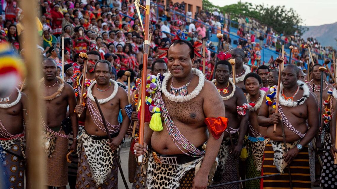 Photo of Swazis in ceremonial dress, with King Mswati in the center, bare-chested, carrying a ceremonial staff.