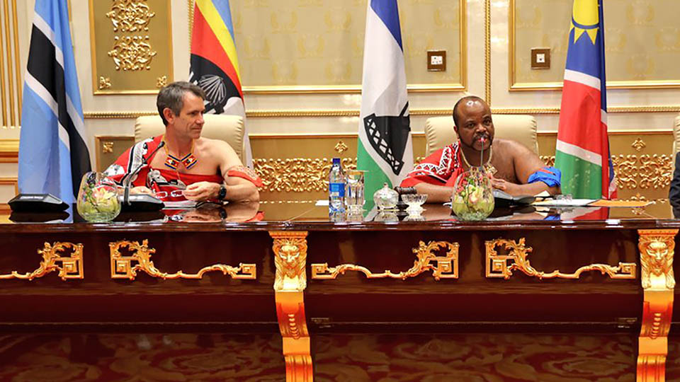 Two men in traditional Swazi dress sit at an elaborate table, with national flags of various African nations in the background.