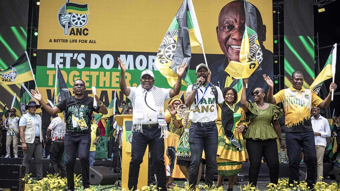 People stand with raised arms, adorned with ANC party colors of yellow, green and black, in front of a large sign that reads ANC, LET'S DO MORE TOGETHER and features the face of President Cyril Ramaphosa.