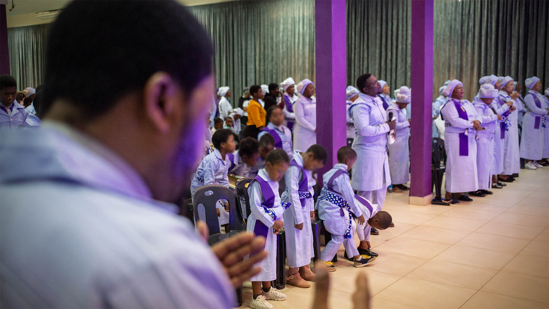 An out-of-focus man in profile in the foreground holds his hands out towards a congregation dressed largely in purple and white robes.