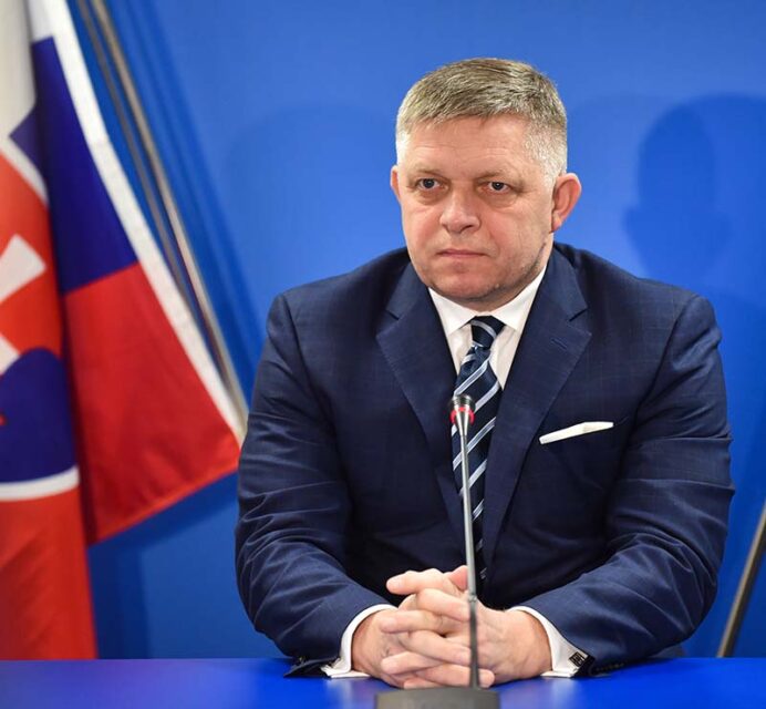Photo of a man in a suit sitting behind a microphone, against a blue backdrop with the corner of the Slovakian flag visible.