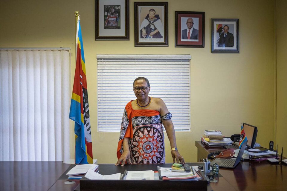 A man in a ceremonial kanga stands in front of a desk beside an Eswatini flag, with photos on a wall behind him depicting Swazi royals and leaders.