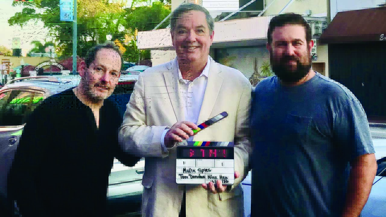 Three men pose on a street, with Thomas Maier in the center holding a film clapperboard.