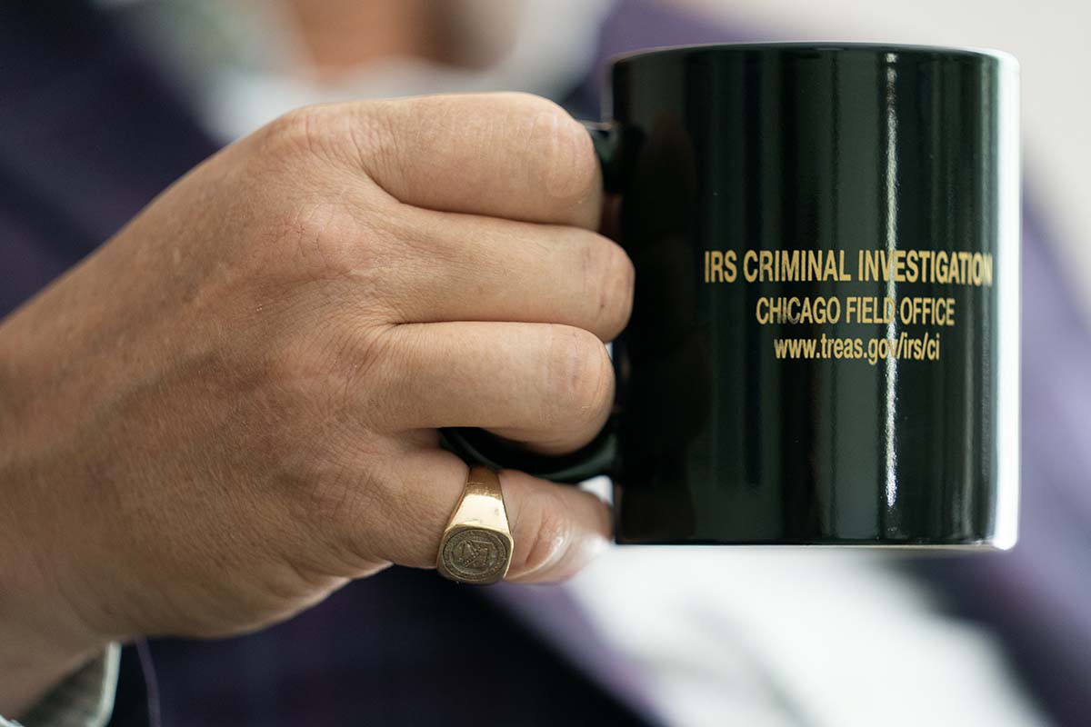 Close-up photo of a hand with a large gold ring on one finger and holding a black mug that reads IRS Criminal Investigation Chicago Field Office.