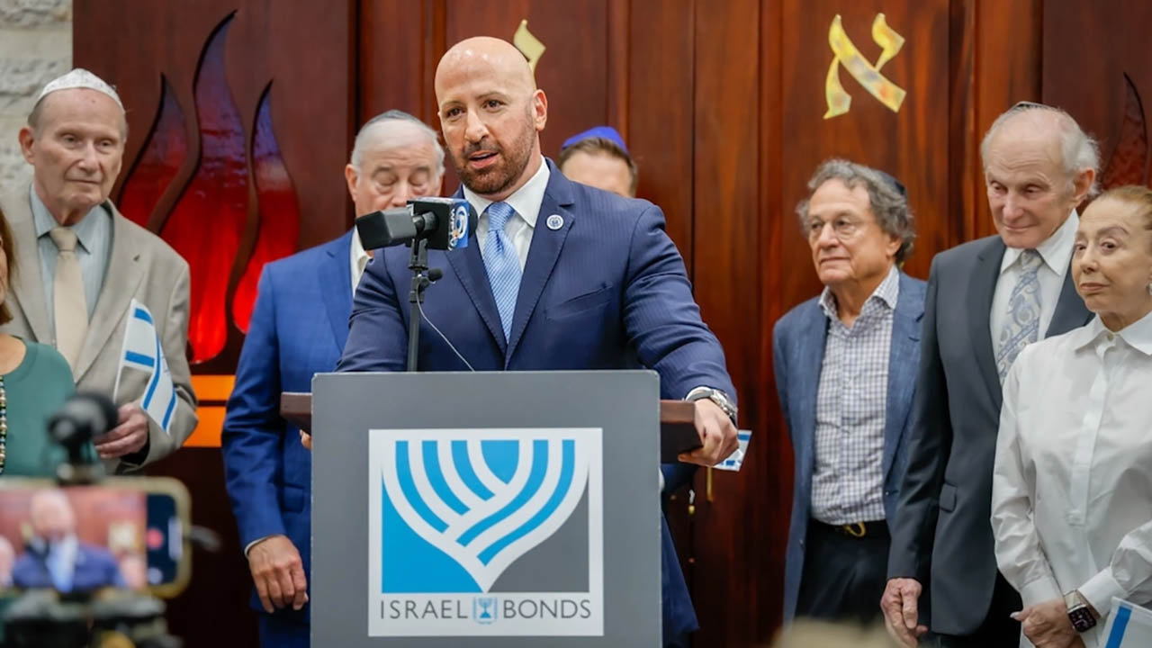 Photo of suited man at a lectern, behind a sign showing the Israeli Bonds logo.