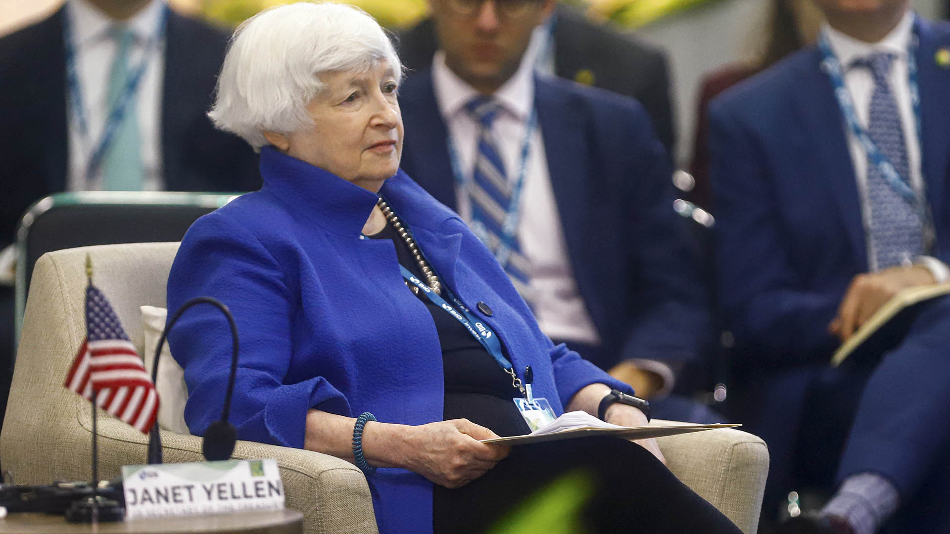 Photo of Janet Yellen sitting in a meeting in front of a nameplate and a small US flag.
