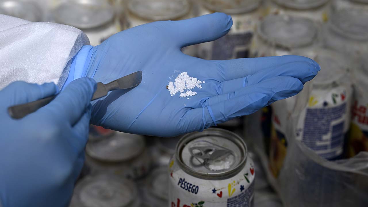 A hand wearing a blue glove shows white powder, with peach juice cans in the background