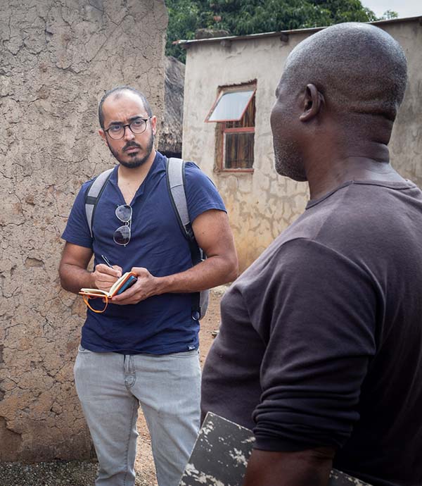 Fotografía que muestra a un periodista con un bloc de notas entrevistando a un hombre parado en medio de un grupo de edificios rurales.