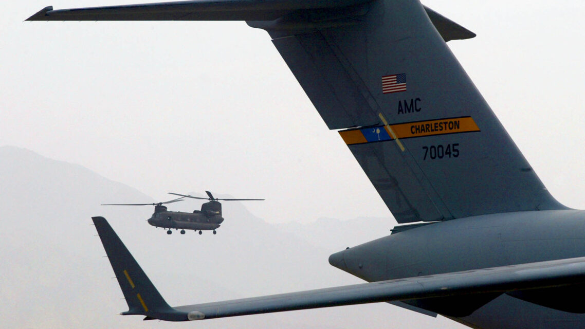 Foreground shows the tail of a military airplane with a small U.S. flag insignia, in the background a Chinook helicopter flies.