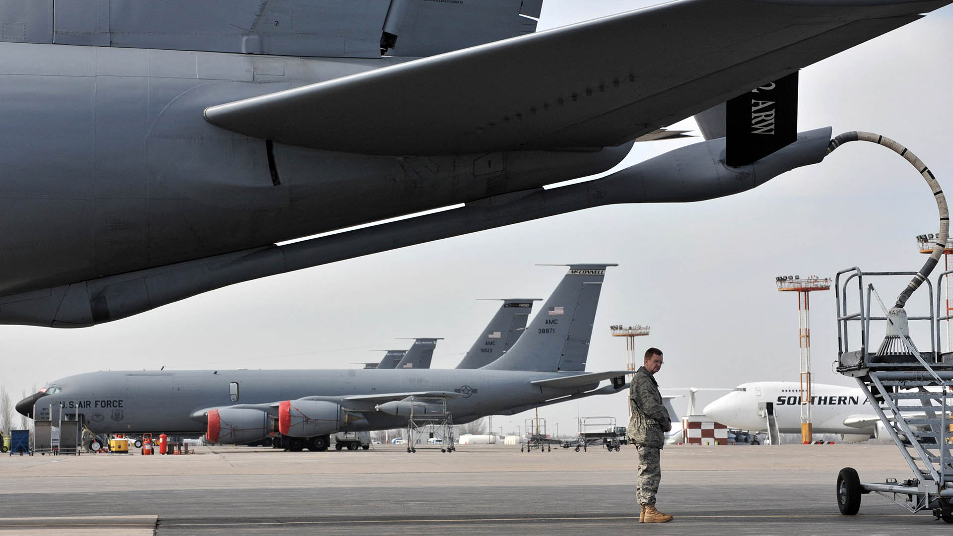 Photo of the tail of a grey plane connected to a fuel hose with a uniformed soldier standing underneath and planes parked in the background at the Manas air base in Bishkek.