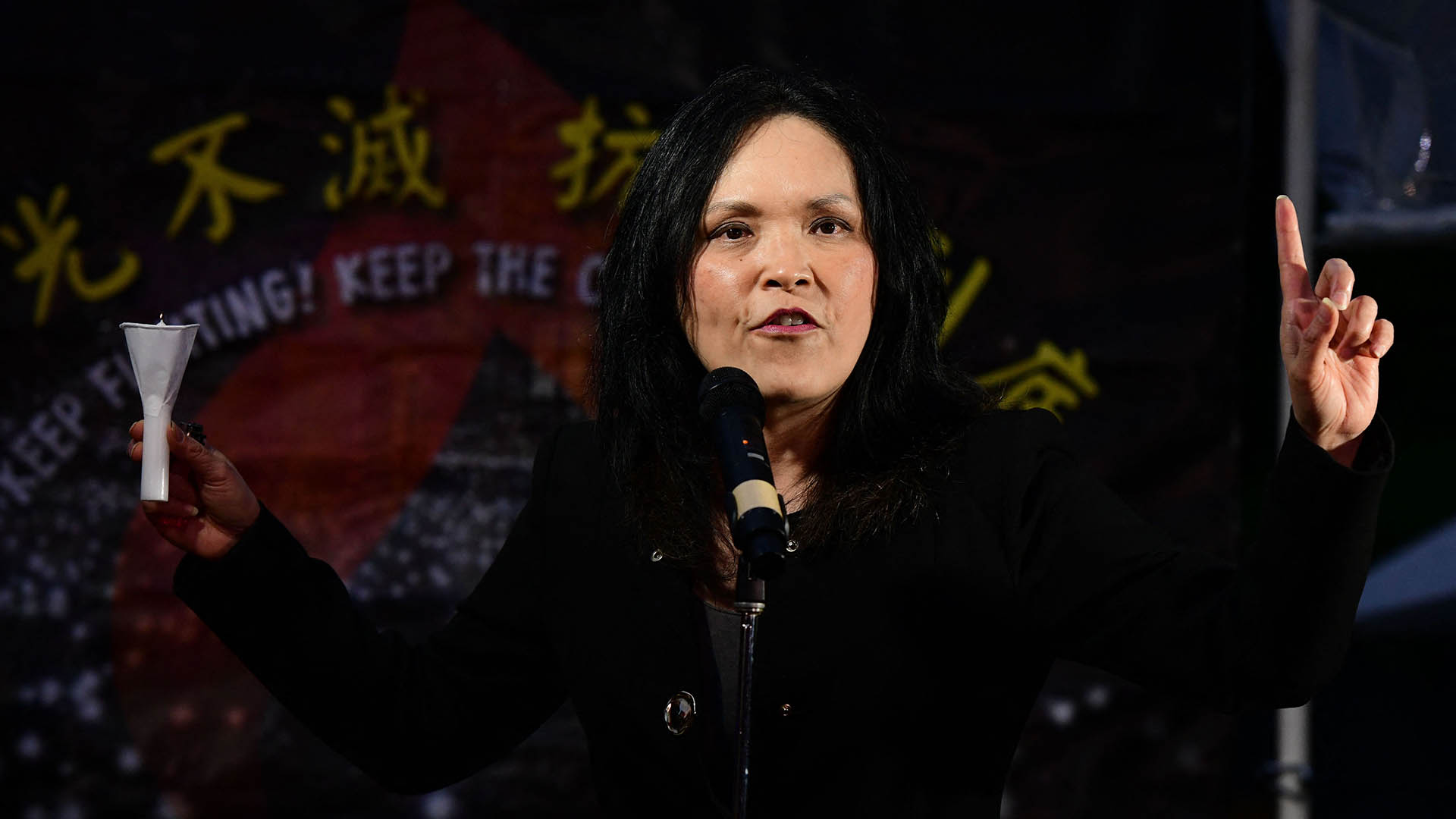 Canadian MP Jenny Kwan gestures and holds a candle in front of a sign with Chinese and English lettering.