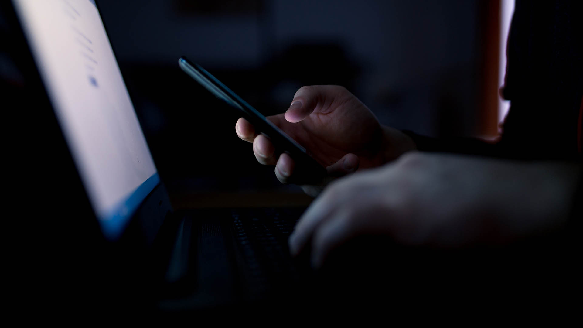 Photo showing hands holding a mobile phone in darkness, lit only by a glowing computer screen.