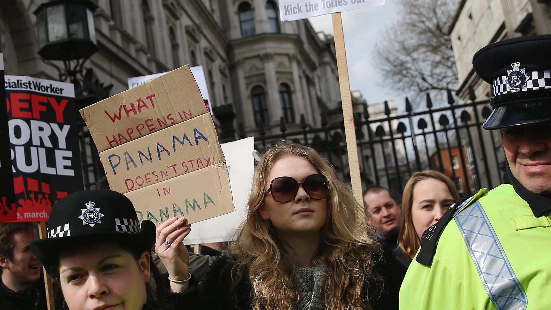 Photo of protesters holding signs behind a police line, including one sign that reads 'What happens in Panama doesn't stay in Panama'