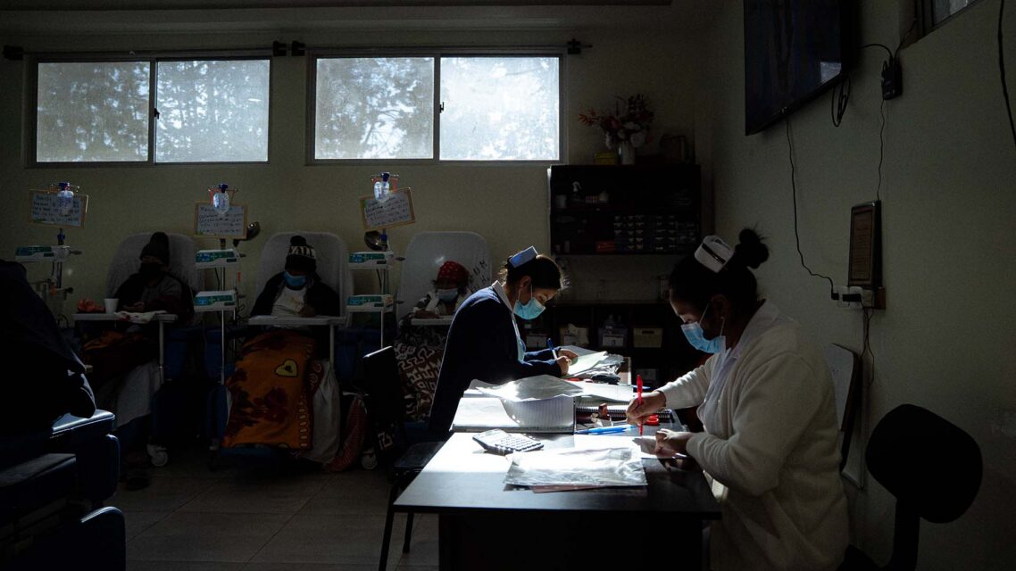 Photo of three patients sitting in chairs with IV drip bags, with nurses doing paperwork in the foreground.