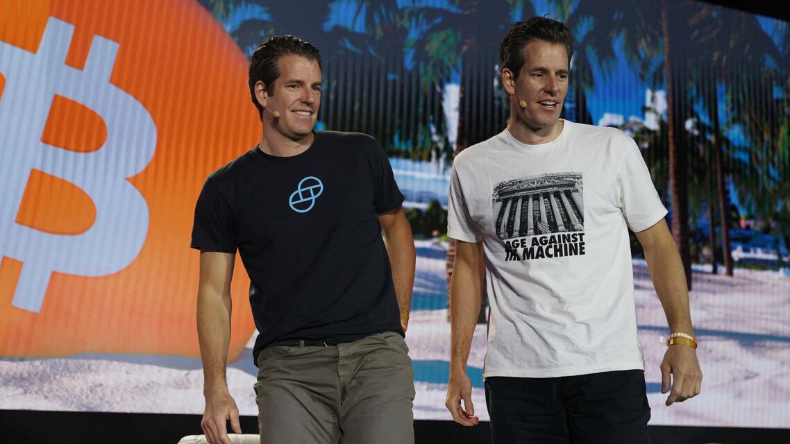 Photo of the Winklevoss twins on a stage in front of a large screen with a bitcoin logo on display.
