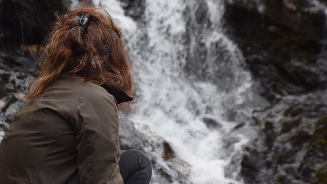 Photo of Helen facing away from the camera, sitting in front of a waterfall.