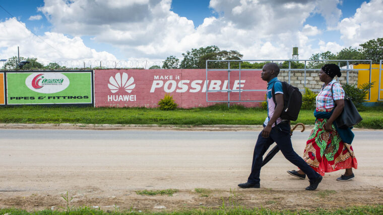 Two people walk past a red billboard for Huawei that says "make it possible." 