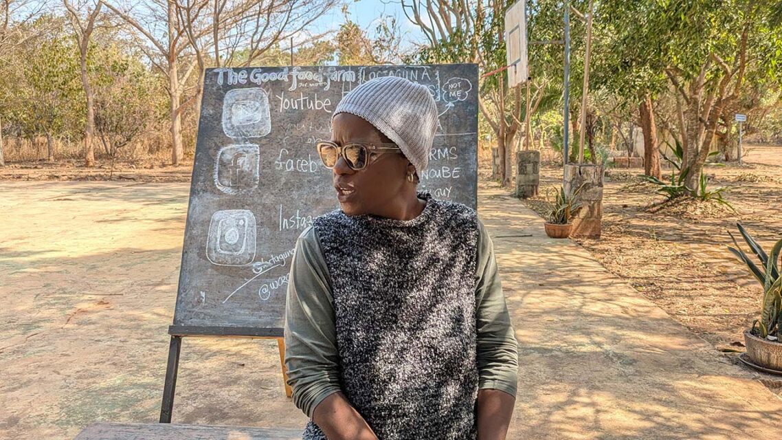 Photo of a woman sitting in front of a chalkboard in an open field on a farm in Zambia.